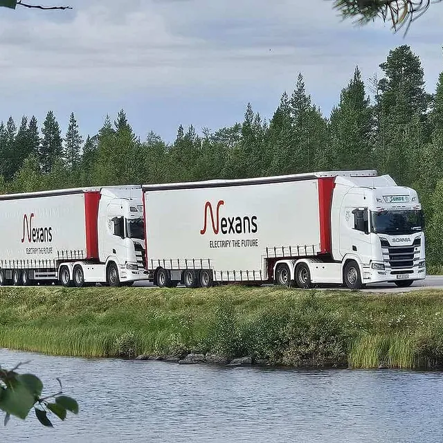 To vogntog kjører langs vei ved elv, omgitt av skog og grønt sommerlandskap.