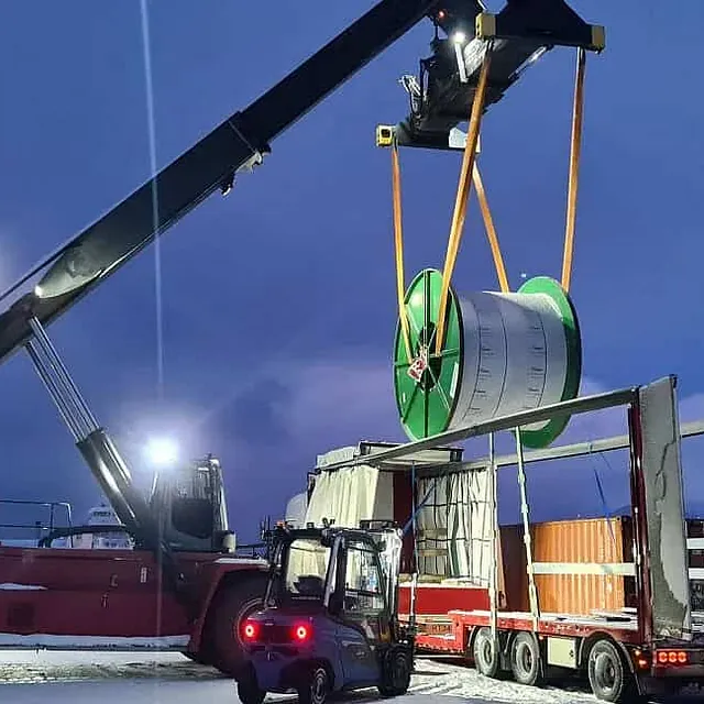 Kran løfter kabeltrommel inn i semitrailer på snødekt terminalområde i vintermørket.