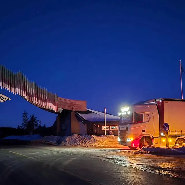 Vogntog står ved Nord-Norge-porten i vintermørket, med opplyst trailer og snødekte omgivelser.