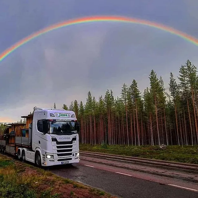 Lastebil med spesialtransport står på landevei under dobbel regnbue ved skogkledd landskap.