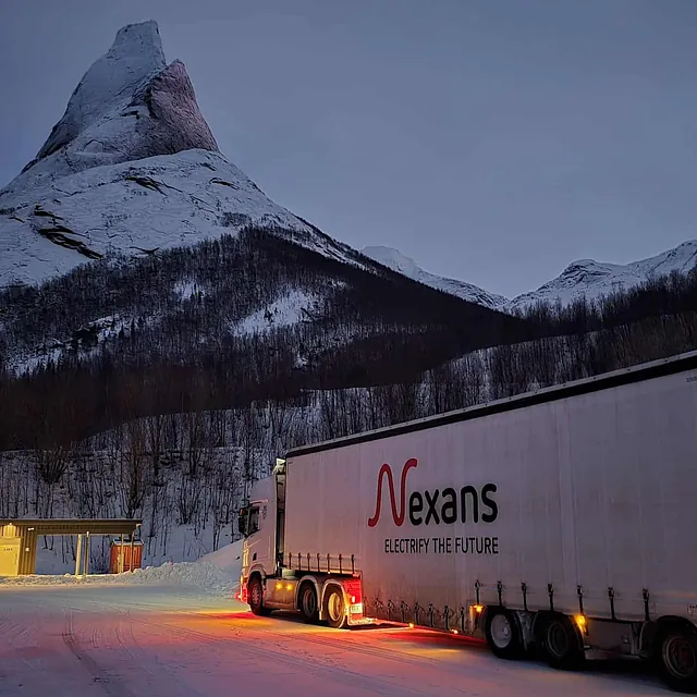 Vogntog parkert i snødekt fjellandskap ved skarp fjelltopp og opplyst rasteplass i skumring.