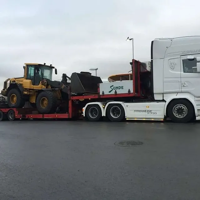 Lastebil transporterer hjullaster på lavbygget henger, parkert på terminalområde i overskyet vær.