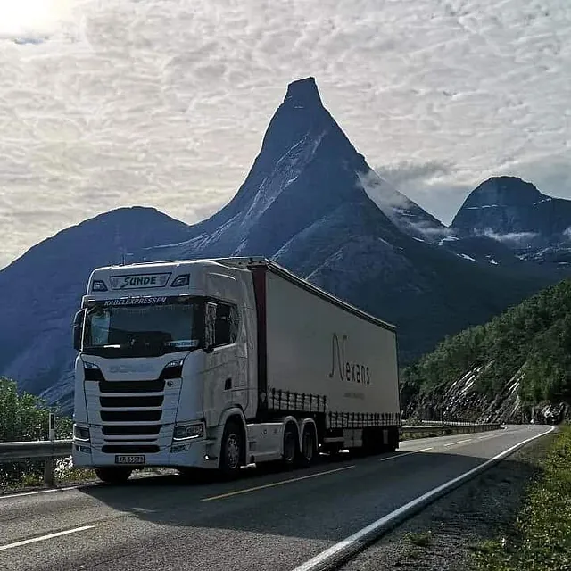 Vogntog kjører langs fjellvei med bratte fjellsider og karakteristiske fjelltopper i bakgrunnen.