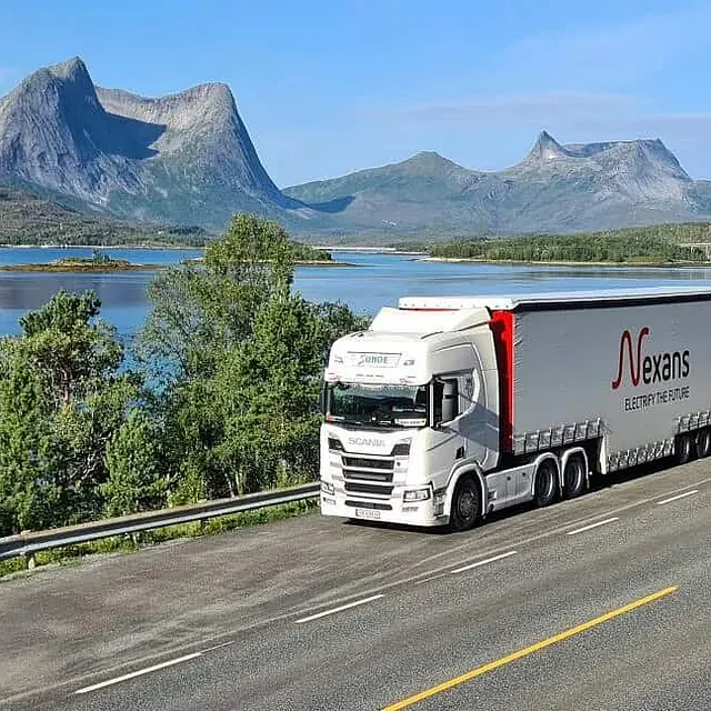 Vogntog kjører langs kystvei med fjord, fjell og spektakulært landskap i bakgrunnen.