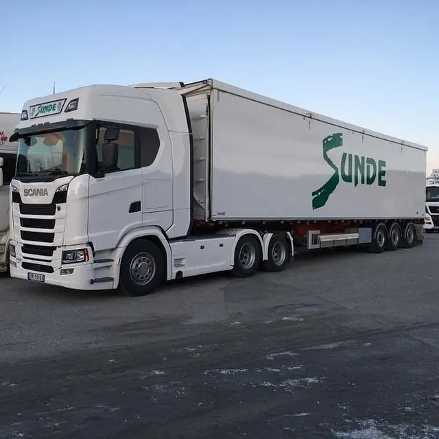 Hvit semitrailer parkert på terminalområde sammen med andre lastebiler i dagslys.
